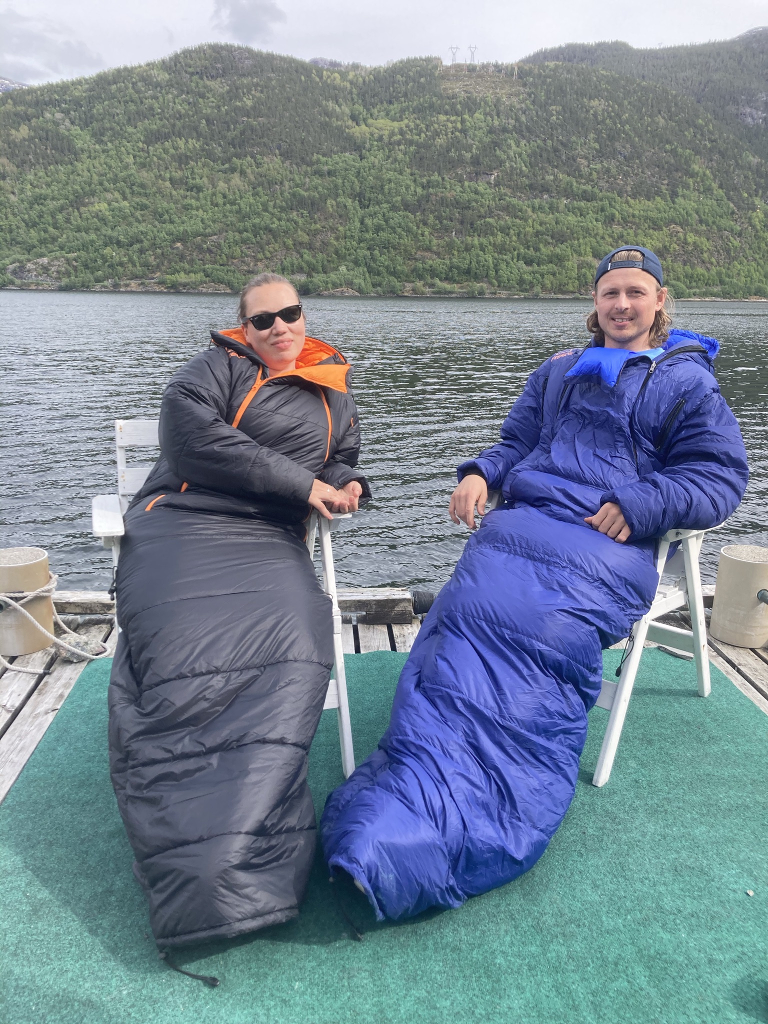 Kvinne og mann med hvert sitt eksemplar av utekos, som sitter på en terrasse ved sjøen.